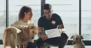 Dog owner pets a tan dog while reviewing paperwork with a caregiver, with another dog resting nearby.