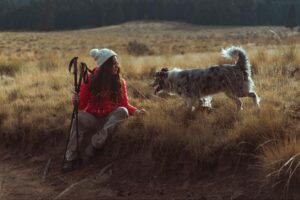 Hiker in a red jacket holding trekking poles rests in a grassy field beside a gray and white dog.